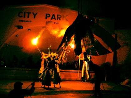 costumed men on stilts waves flaming sticks as a woman dangles in the air at a City Park in Bisbee, AZ