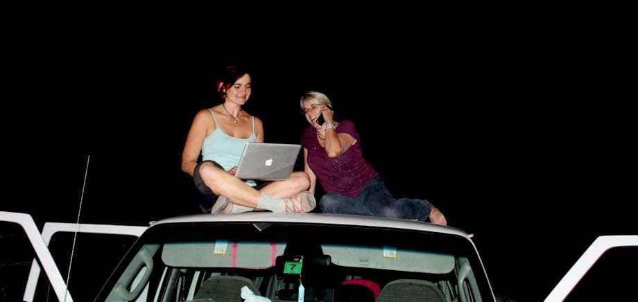 rooftop two women sit on the roof of a van, one talking on her cell phone, the other working on a macbook pro, beneath the night sky