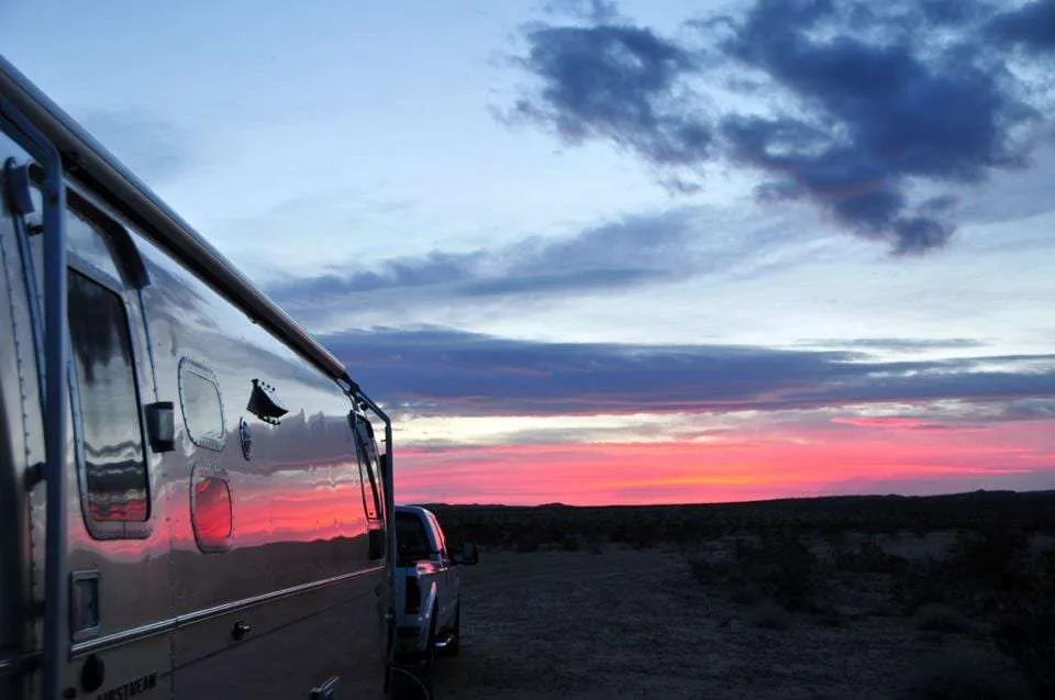 pink divides the silhouette of earth off into the horizon while the evening sky turn blue, all of it reflecting in the side of an aluminum travel trailer