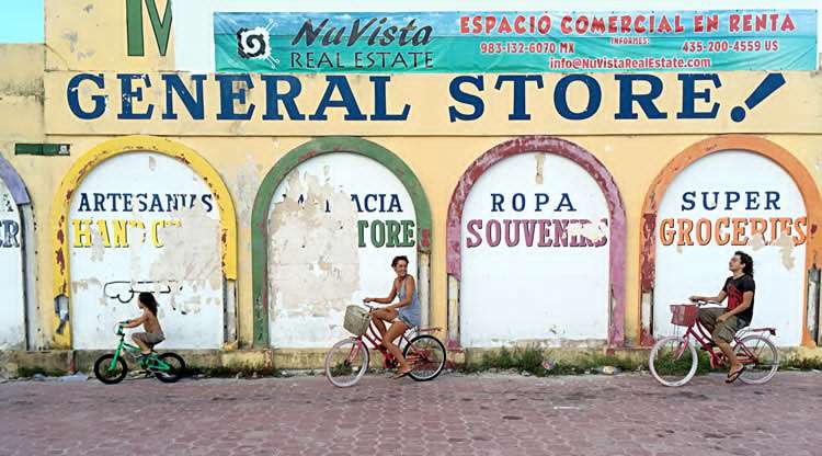 a family on bikes rides in front of a general store in Mahahual, Quintana Roo, Mexico