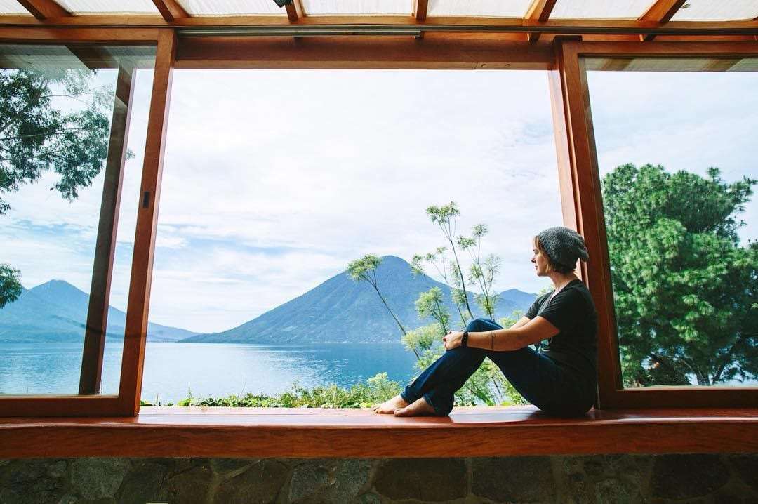 a woman sits in a wooden window, open to the mountains and lake beyond