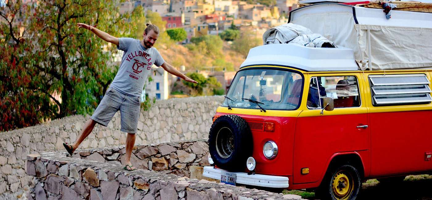 a young man walks a stone wall in Guanajuato, Mexico, his red and yellow VW Bus parked nearby