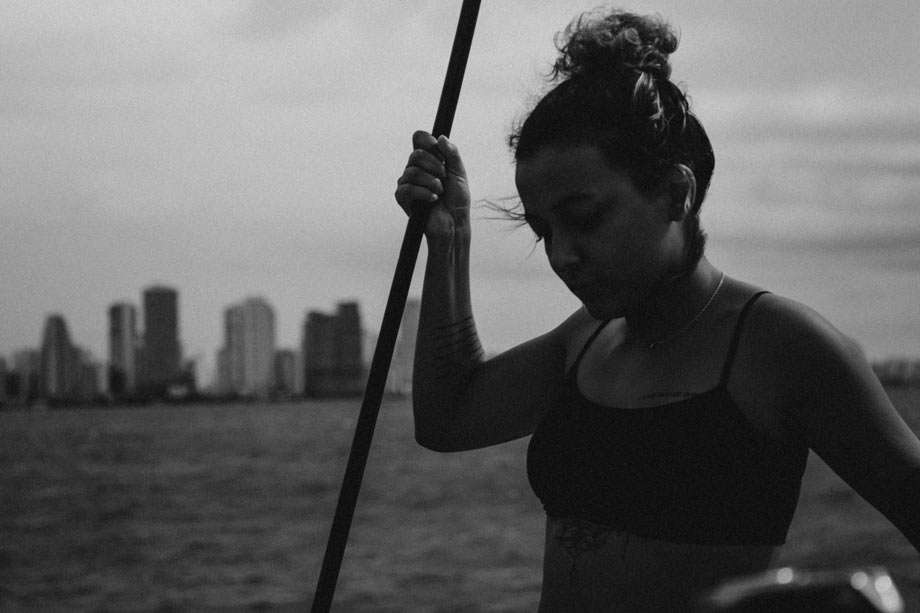 a young woman holds a rope on a sailboat, a city in the background across the water