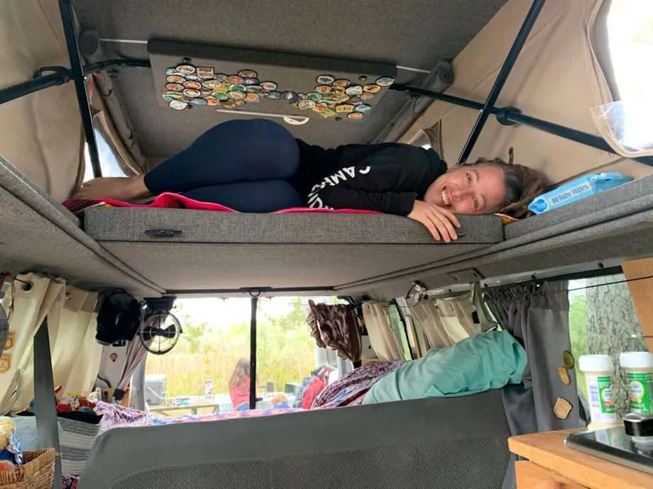 a recently pregnant woman with dreadlocks smiles while laying in the top of a sportsmobile penthouse