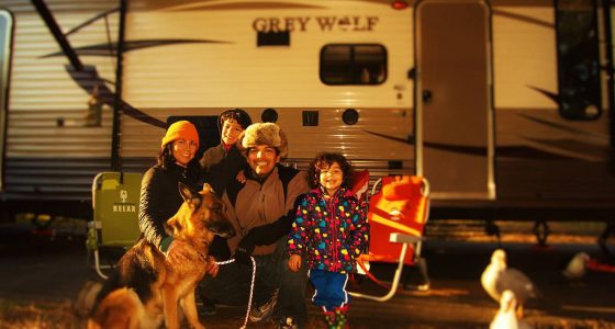 a family of four and their dog sit in camping chairs outside of their Grey Wolf travel trailer