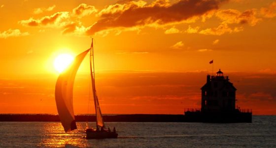 silhouette of a sailboat and a lighthouse against a brilliant burnt orange sky