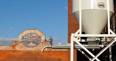 a sign boasting a distressed version of the company logo painted on a brick wall, a new brewing tank in the foreground